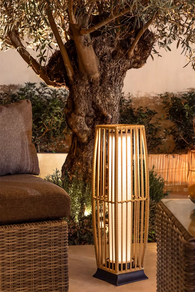 Lámpara de pie solar de diseño en mimbre iluminando un rincón de jardín junto a un árbol, ideal para decoración y ambientación exterior.